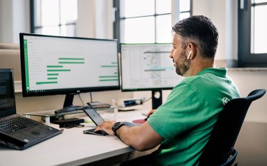 A man in a green t-shirt sits at a desk a looks at a screen.