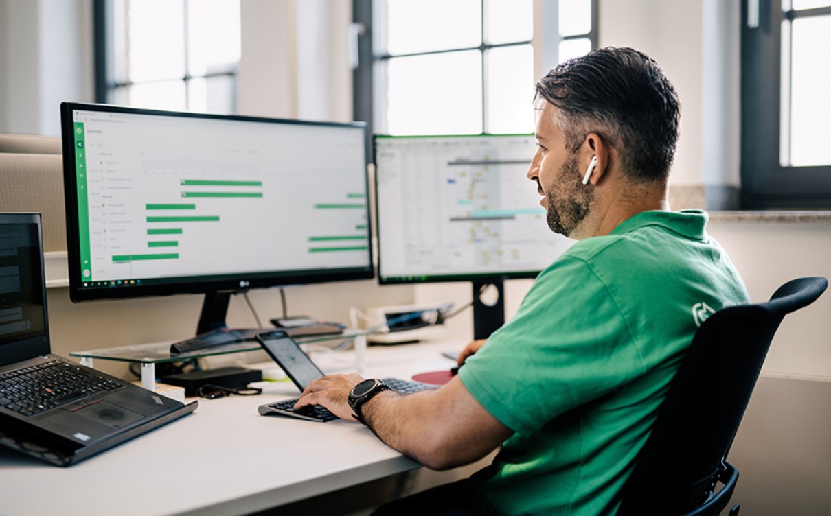 A man in a green t-shirt sits at the desk looking at a screen.