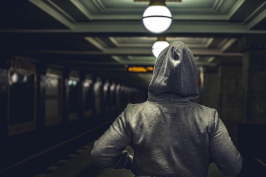 A person in a hoodie walks on a dark train platform.